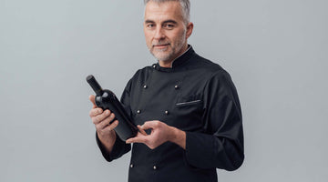 Grauhaariger Sommelie in schwarzer Uniform und Weinflasche in der rechten Hand.