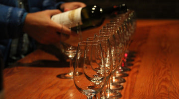 Weingläser aufgereiht auf einem Holztisch, die gerade mit Wein befüllt werden.