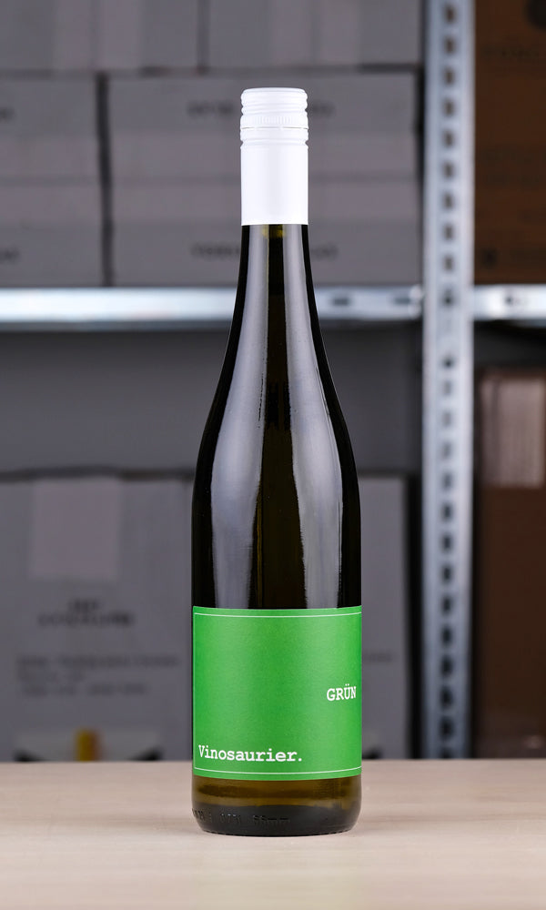 Eine Flasche Vinosaurier Grün vor einem Regal fotografiert. Die Flasche ist aus braunem Glas. Der Verschluss ist weiß. Das Etikett ist grün mit weißer Schrift.