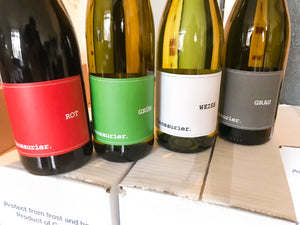 Das Bild zeigt den unteren Teil von vier Weinflaschen, die nebeneinander stehen. V. r. n. l.: Vinosaurier rot, grün, weiß und grau. Sie stehen auf Kartons.