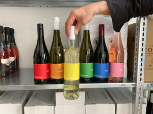 Das Bild zeigt ein Weinregal in der Nahaufnahme. Der Fokus liegt auf sechs Flaschen Vinosaurier Wein unterschiedlicher Farben. Von rechts greift eine Hand ins Bild und holt die Flasche Vinosaurier gelb hervor. 