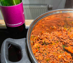 Das Bild zeigt einen Topf von oben. Darin kocht Bolognesesauce. Links dahinter sieht man leicht abgeschnitten eine Flasche Vinosaurier Lila - den Cabernet Sauvignon. 