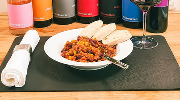In der Mitte steht ein Teller Chili sin Carne, garniert mit drei Scheiben Baguette. Serviert auf einem tiefen Teller. Der Teller steht auf einem Platzdeckchen aus Leder. Rechts hinter dem Teller steht ein Glas Rotwein.