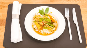 Auf dem Bild ist ein Thai-Curry zu sehen, das auf einem Teller angerichtet wurde. Dieser steht auf einem Platzset aus Leder. Rechts liegt eine Serviette, links Messer und Gabel.