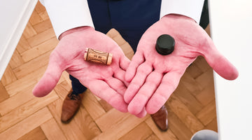 Das Bild zeigt die Handflächen einer Person von oben. In der linken Hand (aus Perspektive des Betrachters) liegt ein Naturkorken, in der rechten Hand ein Schraubverschluss.