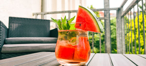 Auf dem Bild ist ein Cocktail zu sehen. Im Glas sind Eiswürfel und Wassermelonenstücke zu erkennen. Der Cocktail ist mit frischer Minze garniert und am Glasrand steckt eine Viertelscheibe Wasseremelone.