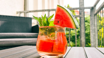 Auf dem Bild ist ein Cocktail zu sehen. Im Glas sind Eiswürfel und Wassermelonenstücke zu erkennen. Der Cocktail ist mit frischer Minze garniert und am Glasrand steckt eine Viertelscheibe Wasseremelone.