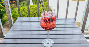 Der Cocktail wurde in einem Weinglas mit Strohhalm angerichtet und steht auf einem Balkontisch. Im Hintergrund sind die Gitter des Balkon und ein Garten zu erkennen. 