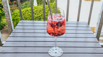Der Cocktail wurde in einem Weinglas mit Strohhalm angerichtet und steht auf einem Balkontisch. Im Hintergrund sind die Gitter des Balkon und ein Garten zu erkennen. 
