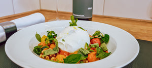 Das Bild zeigt einen Teller mit Burrata im Kirschtomatenbett auf einem Platzdeckchen aus Leder. Dahinter ist leicht verschwommen eine Flasche Vinosaurier grau zu erkennen.