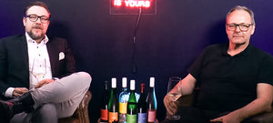 Thomas Coucoulis sitzt links im Bild, Stefan Maas rechts. Beide sind dunkel gekleidet. Sie sitzen vor einer dunkel Kulisse. Zwischen ihnen steht ein Tisch mit allen Vinosaurier Weinen.