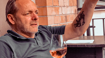 Auf dem Bild ist Stefan Maas auf einem Balkon zu sehen. In der rechten Hand hält er ein Glas Roséwein. Den linken Arm hebt er gerade zum Gestikulieren an. Auf seinem linken Arm ist ein Ankertattoo zu sehen. 
