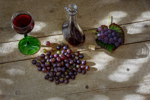 Das Bild zeigt zwei Haufen rote Trauben, ein Glas mit Rotwein gefüllt und einen Dekantiere mit Rotwein. Sie stehen auf einem hölzernen Untergrund.
