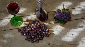 Das Bild zeigt zwei Haufen rote Trauben, ein Glas mit Rotwein gefüllt und einen Dekantiere mit Rotwein. Sie stehen auf einem hölzernen Untergrund.