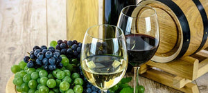Stillleben. Ein Glas Rotwein, ein Glas Weißwein. Daneben rote und grüne Trauben auf einem Holzbrett. Im Hintergrund ist der vordere Teil eines kleinen Weinfasses zu sehen.