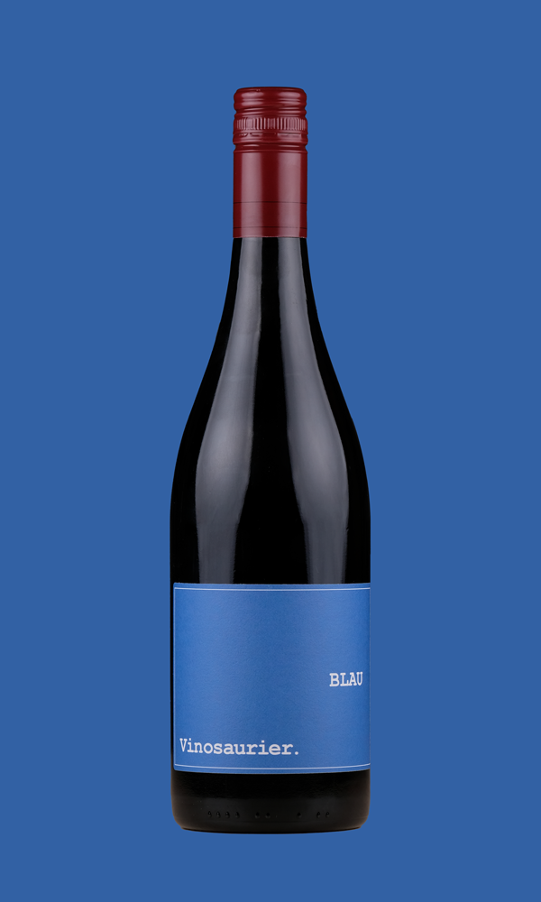 Eine Flasche Vinosaurier blau vor einem blauen Hintergrund. Die Färbung der Flasche ist aufgrund der dunklen Farbe des Rotweins nicht zu kennen. Der Verschluss ist dunkelrot, das Etikett ist blau. 