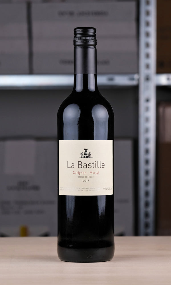 Eine Flasche La Bastille Rouge vor einem Regal fotografiert. Die Färbung der Flasche ist wegen der dunklen Farbe des Weins nicht zu erkennen, das Etikett ist weiß. Darauf steht mit schwarzer Schrift: "La Bastille". Darunter in dunkelroter Schrift: "Carignan - Merlot"