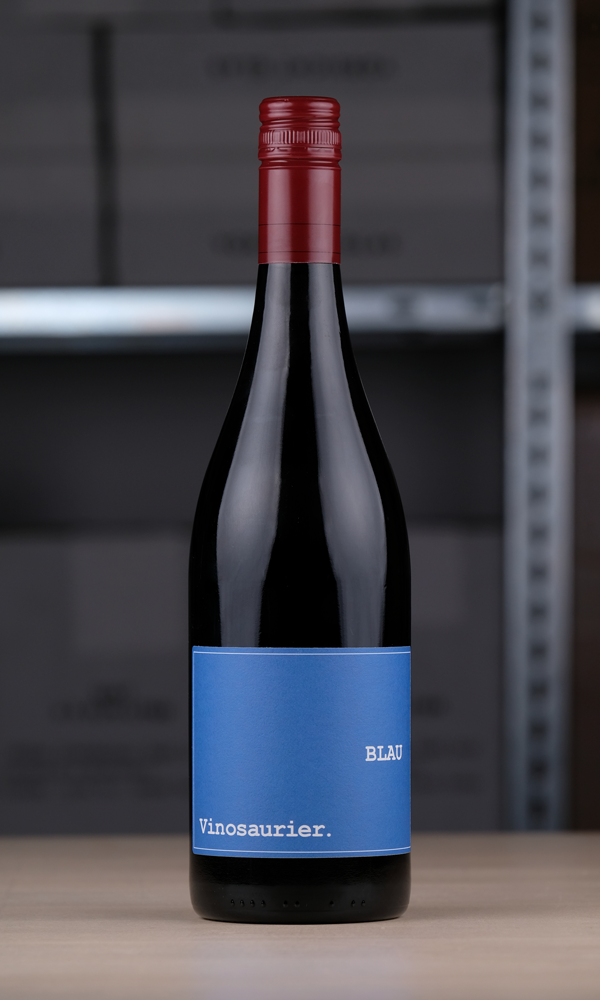 Eine Flasche Vinosaurier blau vor einem Regal fotografiert. Die Färbung der Flasche ist aufgrund der dunklen Farbe des Rotweins nicht zu kennen. Der Verschluss ist dunkelrot, das Etikett ist blau. 
