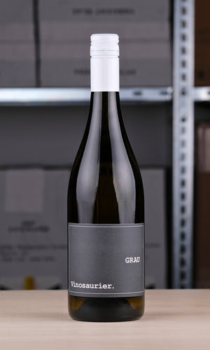 Eine Flasche Vinosaurier Grau vor einem Regal fotografiert. Die Flasche ist aus braunem Glas. Der Verschluss ist weiß. Das Etikett ist grau mit weißer Schrift.