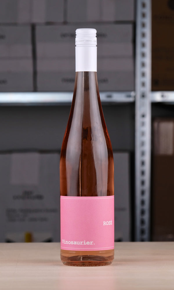 Eine Flasche Vinosaurier rosé vor einem Regal fotografiert. Die Flasche ist durchsichtig, sodass der Roséwein direkt zu sehen ist. Das Etikett ist rosa mit weißer Schrift. Auch der Verschluss ist weiß.