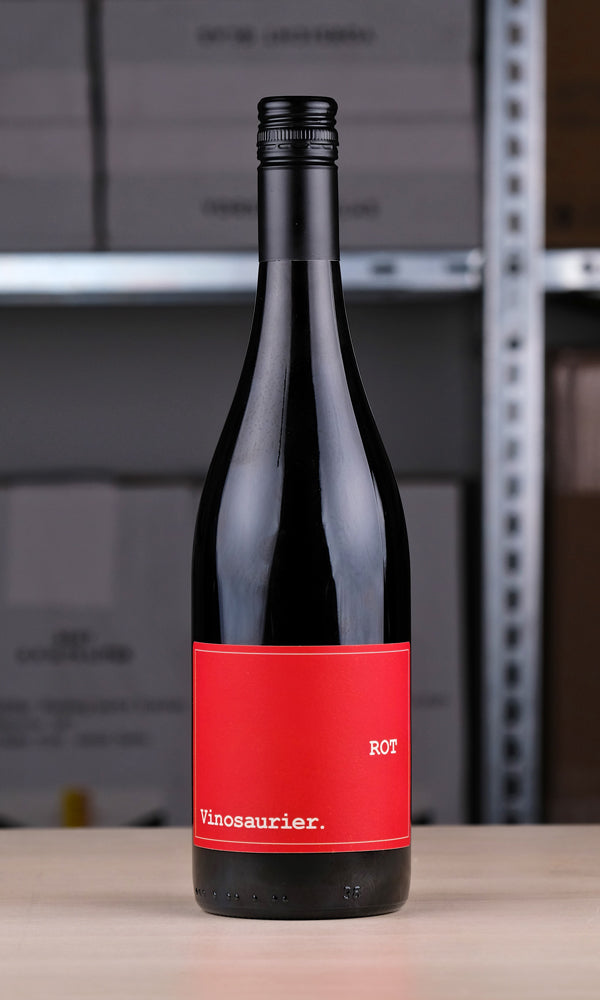 Eine Flasche Vinosaurier rot vor einem Regal abgelichtet. Die Färbung der Flasche ist aufgrund der dunklen Farbe des Rotweins nicht zu kennen. Der Verschluss ist schwarz das Etikett ist rot mit weißer Schrift. 